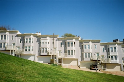 A row of modern townhouses with uniform architectural design stands on a sloped piece of land. Each unit has multiple floors with large, multi-pane windows and balconies. The sky is clear and blue, and there is a car parked in the driveway.