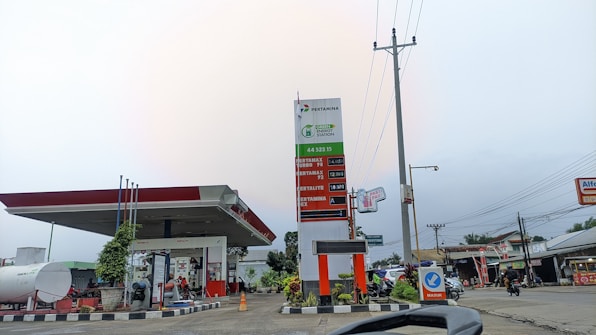 A gas station with a large sign displaying fuel prices and a canopy over the pumps. There are several signs and advertisements around, including one for a convenience store. The sky is overcast, and there are vehicles on the adjacent road.