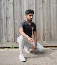 A person is kneeling on one knee on a concrete surface in front of a wooden fence. They are wearing a dark-colored t-shirt, light gray sweatpants, and white sneakers.