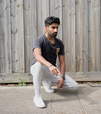 A person is kneeling on one knee on a concrete surface in front of a wooden fence. They are wearing a dark-colored t-shirt, light gray sweatpants, and white sneakers.