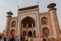 An ornate, grand gateway made of red sandstone with intricate white marble inlay designs. The architectural style is Mughal, featuring large arched entrances and decorative minarets. A crowd of diverse people is gathered in front, some walking towards the entrance, while others are standing and taking photographs. The sky is overcast, creating a subdued lighting over the scene.