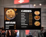 A menu board displaying various dishes under the categories 'Biryani' and 'Curries' with corresponding prices. The left side features an image of biryani, while the right shows images of different curries. People are visible in the blurred background.