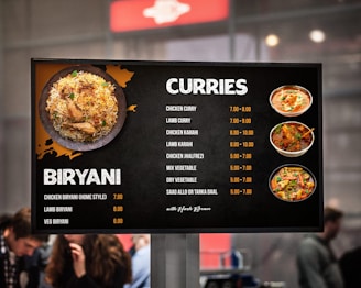 A menu board displaying various dishes under the categories 'Biryani' and 'Curries' with corresponding prices. The left side features an image of biryani, while the right shows images of different curries. People are visible in the blurred background.