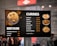 A menu board displaying various dishes under the categories 'Biryani' and 'Curries' with corresponding prices. The left side features an image of biryani, while the right shows images of different curries. People are visible in the blurred background.