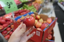 A person holds a red apple in their hand in a grocery store. The background includes a display of apples in a cardboard box and shelves stocked with various fruits. A price sign is visible, indicating a promotion.