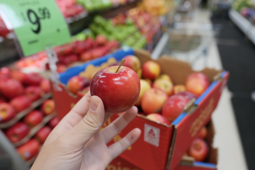 A person holds a red apple in their hand in a grocery store. The background includes a display of apples in a cardboard box and shelves stocked with various fruits. A price sign is visible, indicating a promotion.
