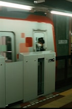A modern train station platform features a sleek, red and white train speeding by. A platform gate is closed, with a sign displaying platform numbers. A person in uniform is visible inside the train, wearing a mask.