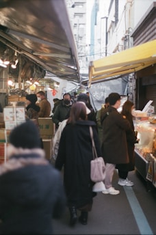 A bustling street market with people walking and shopping. The stalls are covered with tarpaulins, and the scene is filled with various goods and food items. People are dressed in winter clothing, indicating a cold day. The atmosphere is lively, with groups interacting and browsing the market offerings.