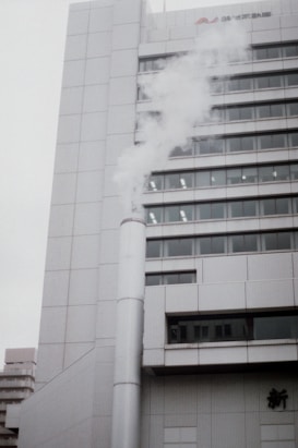 A tall, modern building with a white, tiled exterior emits a large plume of steam or smoke from a pipe attached to its side. The building features multiple windows arranged in a grid pattern, and there is a small amount of Japanese text visible on the upper portion.