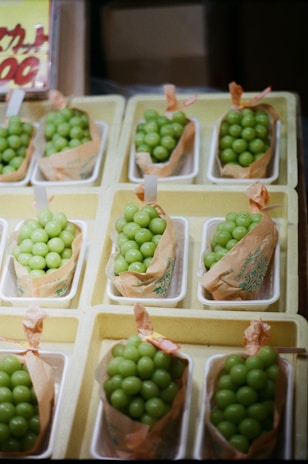 Several containers filled with bright green grapes are neatly arranged in a grid pattern. Each container is lined with a brown paper wrap, and they are placed inside shallow white trays. A partially visible signage with text can be seen in the background.