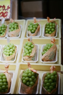 Several containers filled with bright green grapes are neatly arranged in a grid pattern. Each container is lined with a brown paper wrap, and they are placed inside shallow white trays. A partially visible signage with text can be seen in the background.