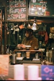 A cozy coffee shop setting features a barista preparing drinks. The scene is framed with warm lighting and handwritten chalk menus hanging above. Various coffee-making equipment and disposable cups are visible, suggesting a focus on artisanal coffee preparation.