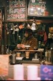 A cozy coffee shop setting features a barista preparing drinks. The scene is framed with warm lighting and handwritten chalk menus hanging above. Various coffee-making equipment and disposable cups are visible, suggesting a focus on artisanal coffee preparation.