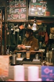 A cozy coffee shop setting features a barista preparing drinks. The scene is framed with warm lighting and handwritten chalk menus hanging above. Various coffee-making equipment and disposable cups are visible, suggesting a focus on artisanal coffee preparation.