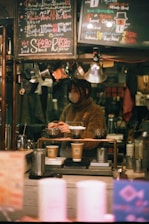 A cozy coffee shop setting features a barista preparing drinks. The scene is framed with warm lighting and handwritten chalk menus hanging above. Various coffee-making equipment and disposable cups are visible, suggesting a focus on artisanal coffee preparation.