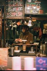 A cozy coffee shop setting features a barista preparing drinks. The scene is framed with warm lighting and handwritten chalk menus hanging above. Various coffee-making equipment and disposable cups are visible, suggesting a focus on artisanal coffee preparation.
