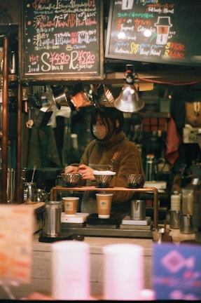 A cozy coffee shop setting features a barista preparing drinks. The scene is framed with warm lighting and handwritten chalk menus hanging above. Various coffee-making equipment and disposable cups are visible, suggesting a focus on artisanal coffee preparation.