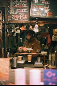 A cozy coffee shop setting features a barista preparing drinks. The scene is framed with warm lighting and handwritten chalk menus hanging above. Various coffee-making equipment and disposable cups are visible, suggesting a focus on artisanal coffee preparation.