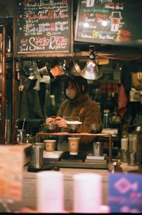 A cozy coffee shop setting features a barista preparing drinks. The scene is framed with warm lighting and handwritten chalk menus hanging above. Various coffee-making equipment and disposable cups are visible, suggesting a focus on artisanal coffee preparation.