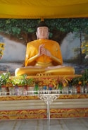 A large Buddha statue is seated in a meditative pose, draped in a golden robe with intricate patterns. The background features a mural of an expansive tree with lush green leaves. In front of the statue, various colorful vases filled with green plants and flowers are arranged on a decorative table.