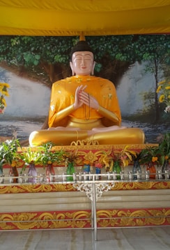 A large Buddha statue is seated in a meditative pose, draped in a golden robe with intricate patterns. The background features a mural of an expansive tree with lush green leaves. In front of the statue, various colorful vases filled with green plants and flowers are arranged on a decorative table.