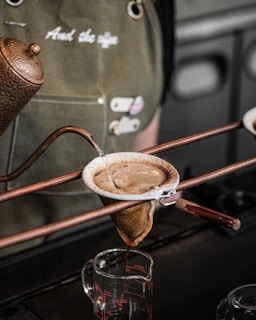 A manual coffee brewing process using a cloth filter held by copper holders. Hot water is poured from a kettle into the filter, which is dripping coffee into a glass measuring cup below. In the background, a person is wearing a green apron with text.