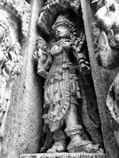 An intricately carved stone sculpture of a deity with detailed ornaments and attire, standing in a niche on an ancient temple wall. The figure appears adorned with elaborate jewelry and a headdress, showcasing fine craftsmanship.