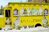 A bright yellow ice cream truck adorned with images of soft-serve ice cream cones featuring different flavors. The truck is parked amidst snow, and there are large illustrations of swirled ice cream cones in white, green, and brown colors on its sides. Menu boards with text and images of ice cream are mounted on the sides of the truck.