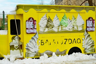 A bright yellow ice cream truck adorned with images of soft-serve ice cream cones featuring different flavors. The truck is parked amidst snow, and there are large illustrations of swirled ice cream cones in white, green, and brown colors on its sides. Menu boards with text and images of ice cream are mounted on the sides of the truck.