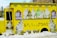 A bright yellow ice cream truck adorned with images of soft-serve ice cream cones featuring different flavors. The truck is parked amidst snow, and there are large illustrations of swirled ice cream cones in white, green, and brown colors on its sides. Menu boards with text and images of ice cream are mounted on the sides of the truck.