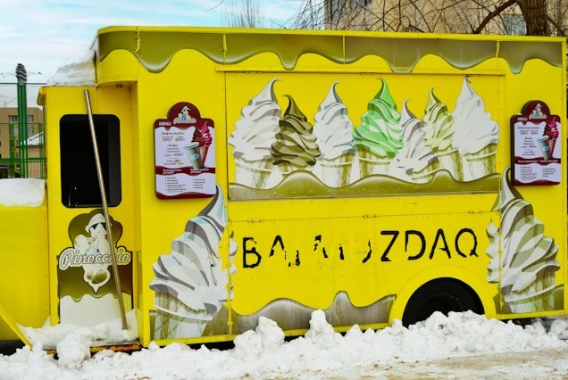 A bright yellow ice cream truck adorned with images of soft-serve ice cream cones featuring different flavors. The truck is parked amidst snow, and there are large illustrations of swirled ice cream cones in white, green, and brown colors on its sides. Menu boards with text and images of ice cream are mounted on the sides of the truck.