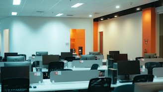 An empty office space with multiple workstations, each equipped with a computer monitor and office chairs. The room is well-lit with overhead fluorescent lights and features white and orange walls.