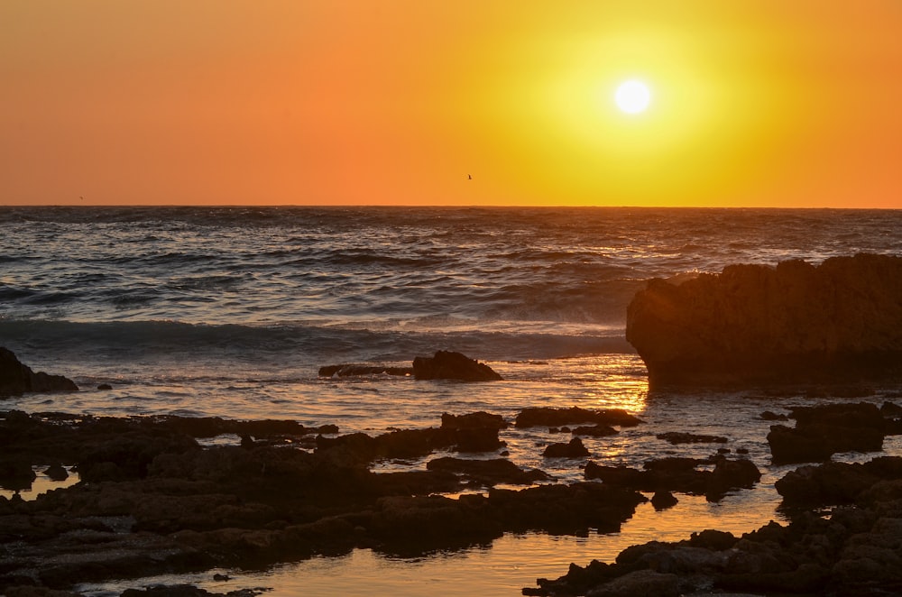 the sun is setting over the ocean with rocks in the foreground