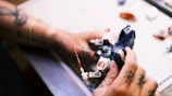 Hands holding and assembling a small robot figurine on a table. The person has tattoos and seems to be focused on the task. The background includes various small parts and tools.