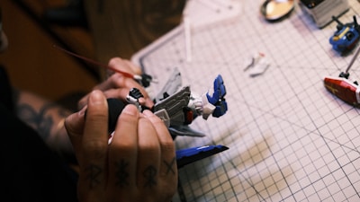 A person is assembling a model robot on a grid-lined cutting mat, with visible hands holding parts of the model. Two disassembled parts lie nearby, and the person's hand has tattoos.