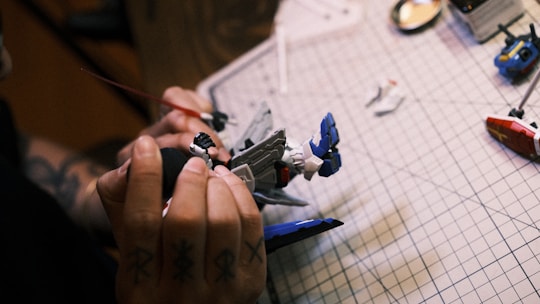 A person is assembling a model robot on a grid-lined cutting mat, with visible hands holding parts of the model. Two disassembled parts lie nearby, and the person's hand has tattoos.