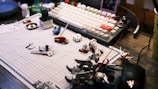 A work desk displaying a variety of model kit parts and tools spread over a grid cutting mat. A mechanical keyboard with white and beige keys is visible in the background, along with various small containers and a mouse. The scene suggests a hobbyist assembling model figures.