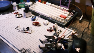 A work desk displaying a variety of model kit parts and tools spread over a grid cutting mat. A mechanical keyboard with white and beige keys is visible in the background, along with various small containers and a mouse. The scene suggests a hobbyist assembling model figures.