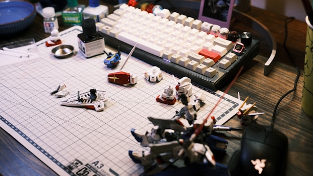 A work desk displaying a variety of model kit parts and tools spread over a grid cutting mat. A mechanical keyboard with white and beige keys is visible in the background, along with various small containers and a mouse. The scene suggests a hobbyist assembling model figures.