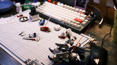 A work desk displaying a variety of model kit parts and tools spread over a grid cutting mat. A mechanical keyboard with white and beige keys is visible in the background, along with various small containers and a mouse. The scene suggests a hobbyist assembling model figures.