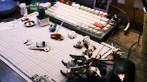 A work desk displaying a variety of model kit parts and tools spread over a grid cutting mat. A mechanical keyboard with white and beige keys is visible in the background, along with various small containers and a mouse. The scene suggests a hobbyist assembling model figures.