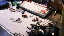 A work desk displaying a variety of model kit parts and tools spread over a grid cutting mat. A mechanical keyboard with white and beige keys is visible in the background, along with various small containers and a mouse. The scene suggests a hobbyist assembling model figures.