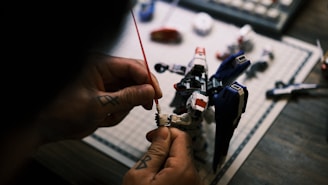 A person is assembling a detailed model robot, focusing on attaching a part to the arm. The model resembles a futuristic mech with red, white, and blue components. The scene is set on a table with other model parts and tools visible.