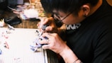 A person with tattoos on their hands is intricately working on a model figurine at a desk. The focus is on their detailed hand movements and the tools they are using. Various parts and tools are scattered on a cutting mat beside them.