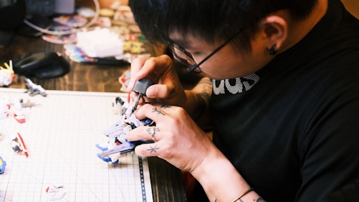 A person with tattoos on their hands is intricately working on a model figurine at a desk. The focus is on their detailed hand movements and the tools they are using. Various parts and tools are scattered on a cutting mat beside them.