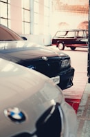 Multiple cars are parked inside a spacious, well-lit garage. The focus is on BMW vehicles, with sleek designs and reflective surfaces. A classic car can be seen in the background, contributing to a vintage atmosphere.