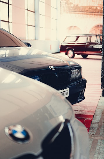 Multiple cars are parked inside a spacious, well-lit garage. The focus is on BMW vehicles, with sleek designs and reflective surfaces. A classic car can be seen in the background, contributing to a vintage atmosphere.