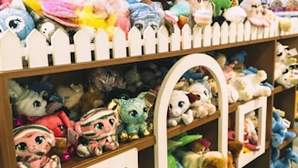 A shelf filled with a variety of plush toys in different colors and designs. The toys are arranged neatly with a decorative white picket fence on top of the shelf. Among the toys are animals with big eyes, including different creatures such as cats and rabbits.