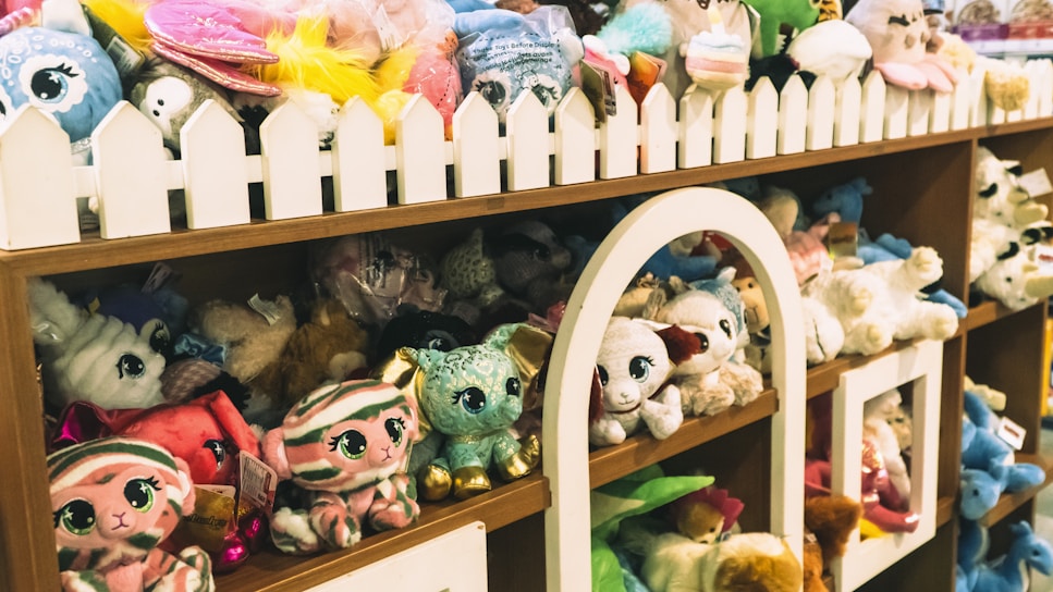 A shelf filled with a variety of plush toys in different colors and designs. The toys are arranged neatly with a decorative white picket fence on top of the shelf. Among the toys are animals with big eyes, including different creatures such as cats and rabbits.