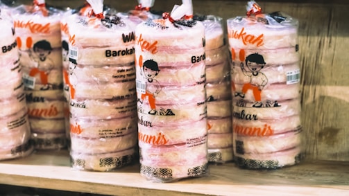 Several stacks of packaged edibles are displayed on a wooden shelf. The packaging has a clear plastic wrap with illustrations of a cartoon character and text in a bright orange color.